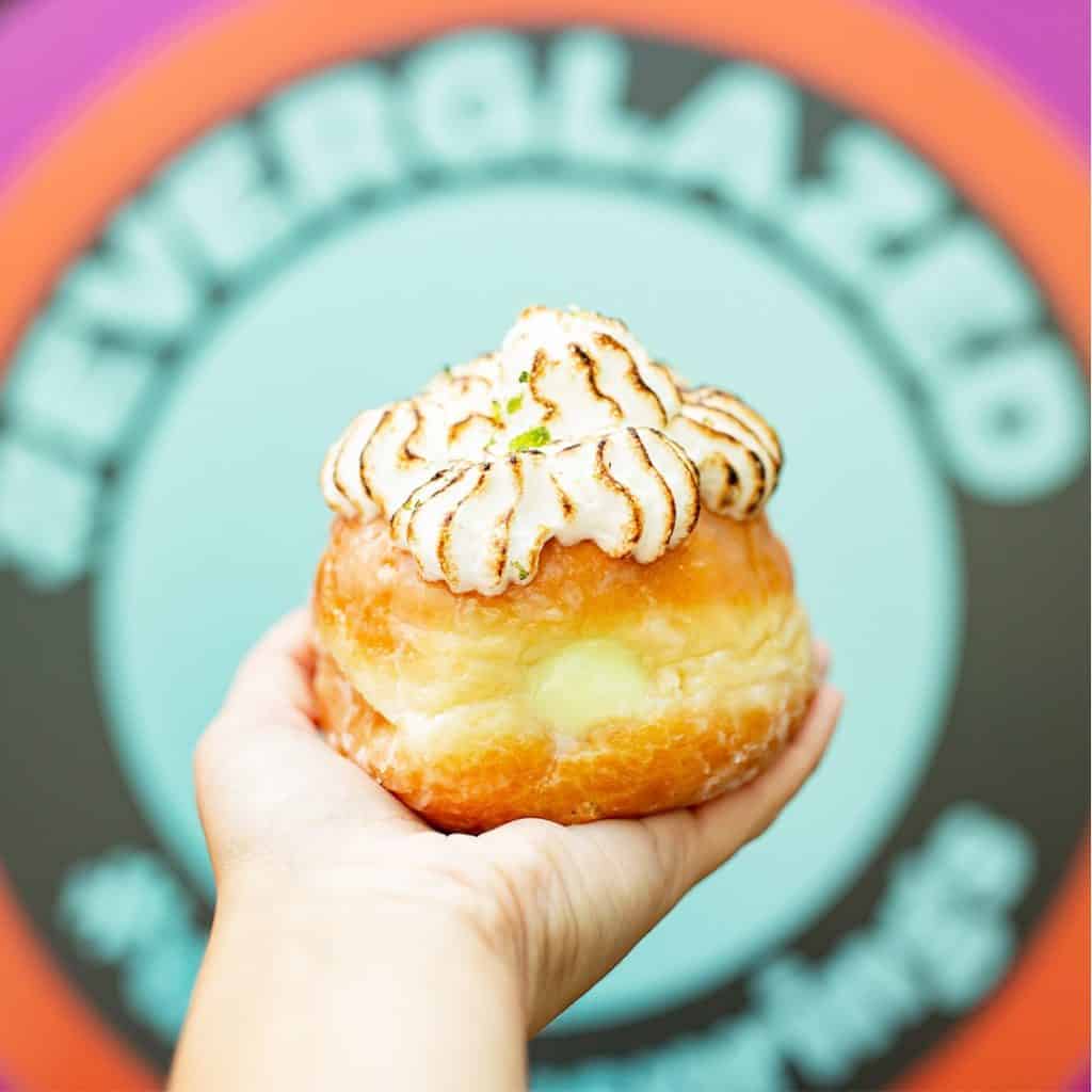 A Key Lime Pie Donut, glazed and crowned with toasted meringue, is held up in front of a vibrant circular sign at Disney Springs—highlighting an irresistible new treat for donut lovers exploring Walt Disney World’s lively shopping and dining district.