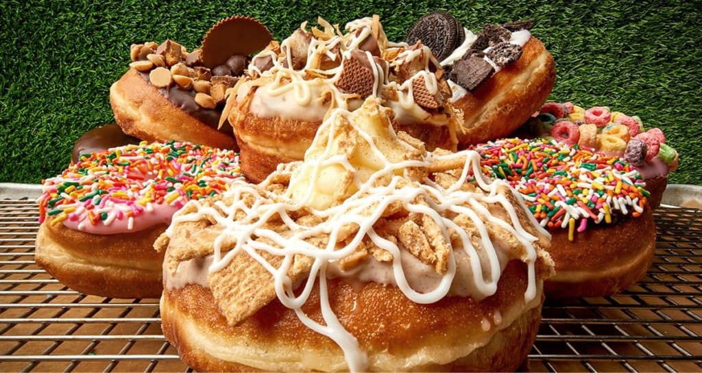 A tempting lineup of gourmet donuts, including our iconic Key Lime Pie Donut, decked out with candy, cookie crumbles, colorful cereal, and sprinkles on a cooling rack—just like the irresistible treats you’ll discover at Disney Springs.