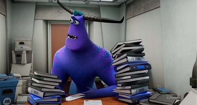 Tylor Tuskmon from Monsters at Work sits at his cluttered Monsters, Inc. office desk, surrounded by towering stacks of books and binders, embodying the lively chaos fans love in Disney’s animated workplace comedies.