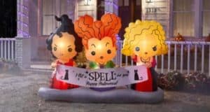 The Sanderson Sisters from Hocus Pocus stand in front of a spellbindingly decorated house at night, holding a “Happy Halloween” banner, surrounded by dazzling Halloween props straight out of your favorite Disney park celebration.