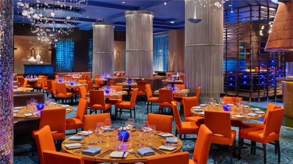 Sleek Disney World restaurant near Crescent Lake featuring vibrant orange chairs, elegantly set tables, striking blue lighting, and towering columns—a chic spot to unwind after a sweet-filled Dessert Crawl at the resort.