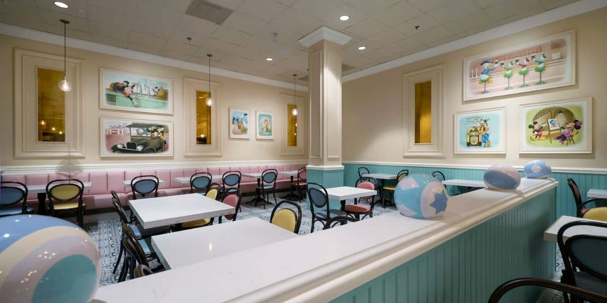 A cozy cafe interior with pastel-colored chairs and white tables. The walls feature various whimsical paintings, and globe-themed balloons are visible in the foreground. The atmosphere is inviting and cheerful.