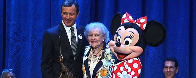 A man, an older woman reminiscent of Disney Legend Betty White, and Minnie Mouse appear together onstage in front of a blue curtain at a Disney event, capturing the whimsical spirit and nostalgic artistry fans recognize from Little Golden Book scenes.
