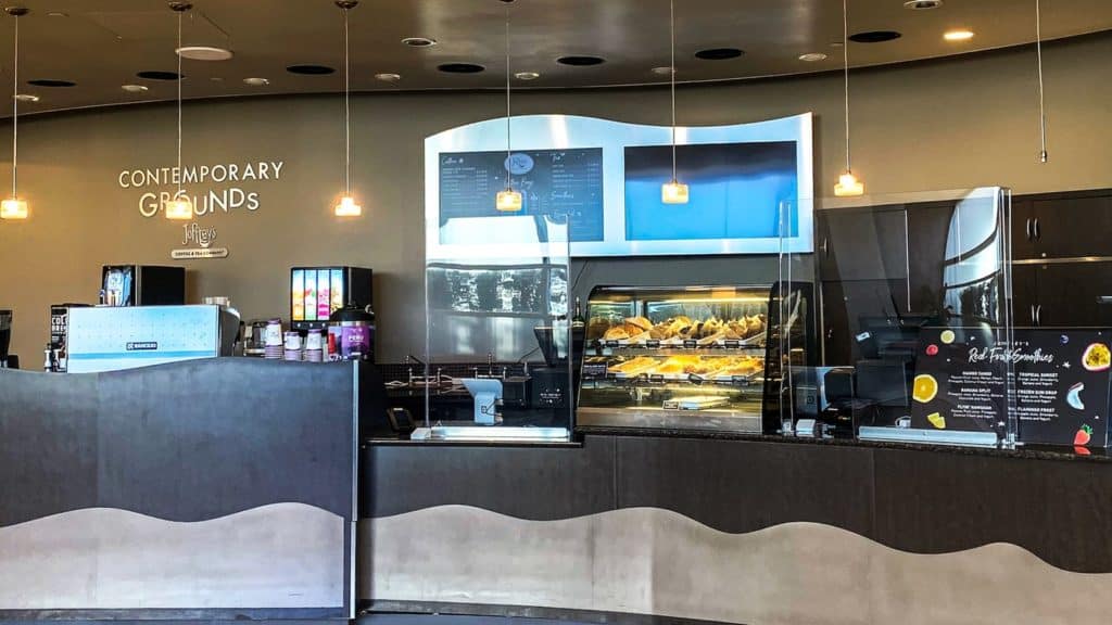 A sleek coffee shop counter at Disney parks featuring a tempting display case of pastries, top-of-the-line coffee machines, and a colorful menu on the wall—capturing the welcoming vibe of Joffrey’s Coffee spots where fans enjoy the magic of mobile ordering to quickly grab their favorite park treats.