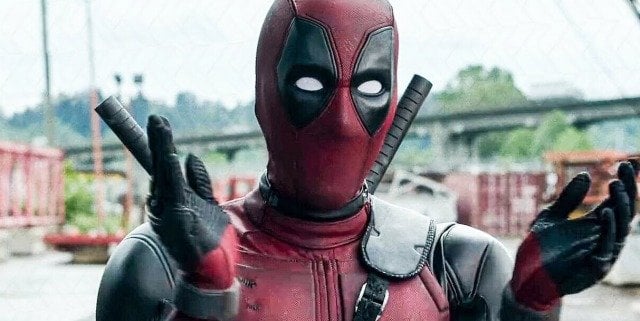 Deadpool, clad in his iconic red and black suit, strikes an animated pose with both hands as he entertains fans on a dynamic bridge set within a lively Marvel-themed attraction, channeling the limitless fun of the Deadpool multiverse.
