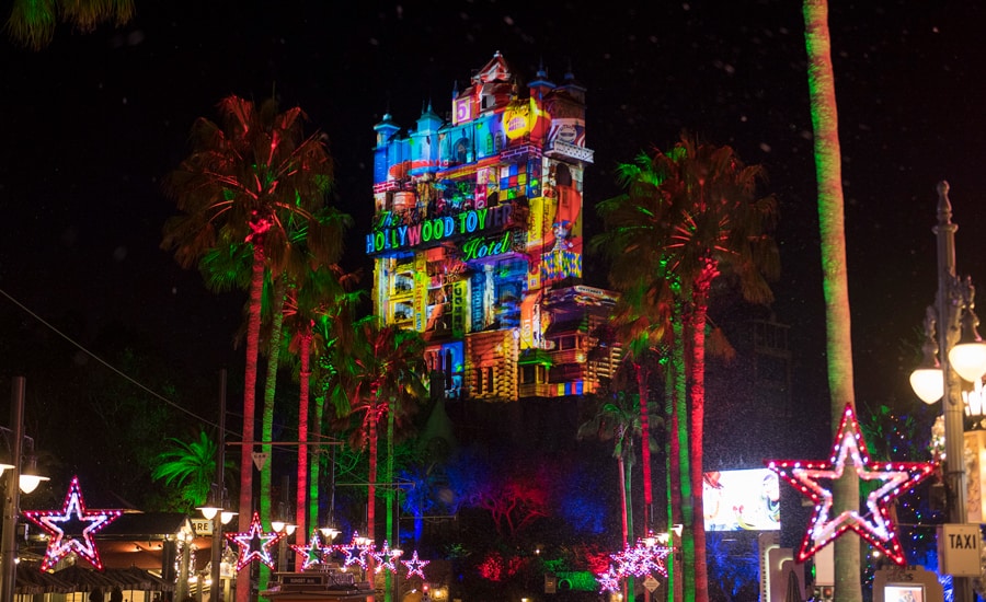 The Hollywood Tower Hotel shines at night at Disney’s Hollywood Studios, illuminated with vibrant holiday lights, surrounded by palm trees and festive glowing star decorations—delivering that unmistakable seasonal magic park guests love.