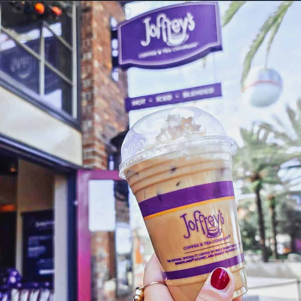 Iced coffee with whipped cream in hand in front of the Joffrey's Coffee shop sign and swaying palm trees, capturing a quintessential Disney snack—now easier than ever to grab with Mobile Ordering at Walt Disney World.