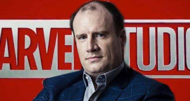 A man in a suit poses in front of a large red and white Marvel Studios logo background.