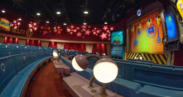 A colorful theater with blue seats and whimsical lamps, featuring a stage with animated screens, star decorations on the ceiling, and a sign reading "Comedy Facility 7" on the back wall.