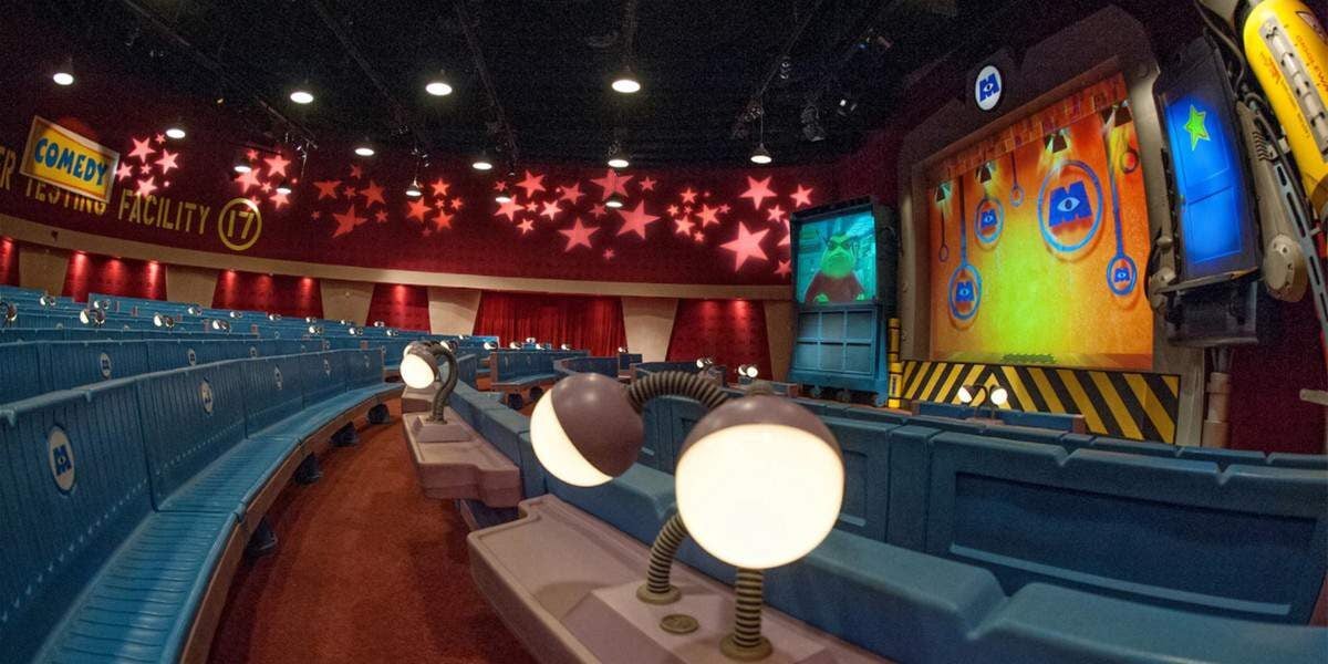 A colorful theater with blue seats and whimsical lamps, featuring a stage with animated screens, star decorations on the ceiling, and a sign reading "Comedy Facility 7" on the back wall.