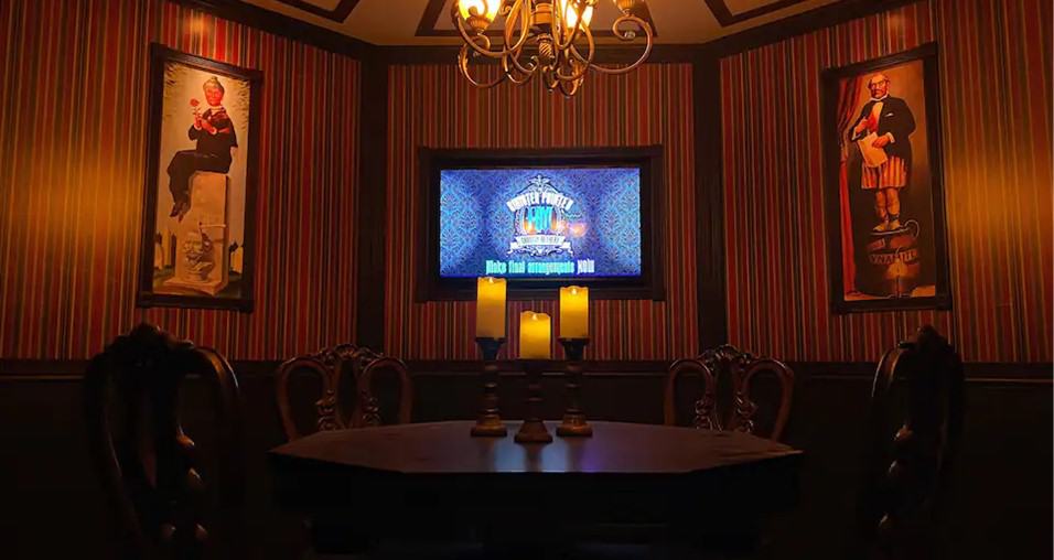 Dimly lit room inside the Haunted Mansion, featuring a round table, vintage chairs, flickering candles, two eerie portraits, and a screen against striped walls—inviting fans to experience the iconic ghostly ambiance themselves.