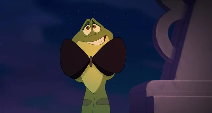 Prince Naveen as a lively green frog beaming shyly, clutching a large black bow tie to his chest under the nighttime theme park lights, channeling his signature charm for fans.