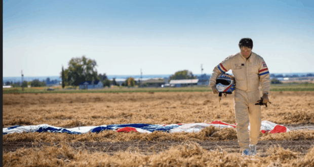 A stunt performer in a flightsuit strides across an open field beneath a clear sky, helmet in hand and parachute nearby—capturing the real-life excitement behind the jaw-dropping stunts showcased in Disney+’s "Stuntman" documentary trailer.