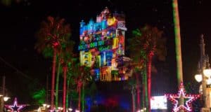 The Hollywood Tower Hotel at Disney’s Hollywood Studios transforms into a dazzling nighttime spectacle during the 2021 holiday season, with vivid holiday projections illuminating the facade, shimmering star lights overhead, and festive palm trees lining the scene—creating a magical atmosphere for park guests.