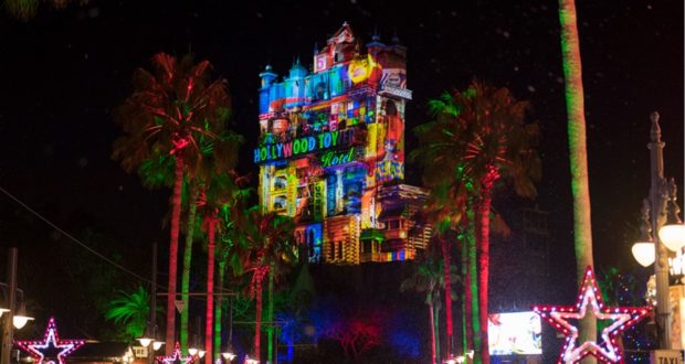The Hollywood Tower Hotel at Disney’s Hollywood Studios transforms into a dazzling nighttime spectacle during the 2021 holiday season, with vivid holiday projections illuminating the facade, shimmering star lights overhead, and festive palm trees lining the scene—creating a magical atmosphere for park guests.
