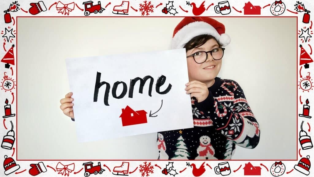 Young guest in a festive sweater and Santa hat poses with a "home" sign featuring a red house, framed by holiday decorations, channeling the excitement for the new Home Alone attraction this season.