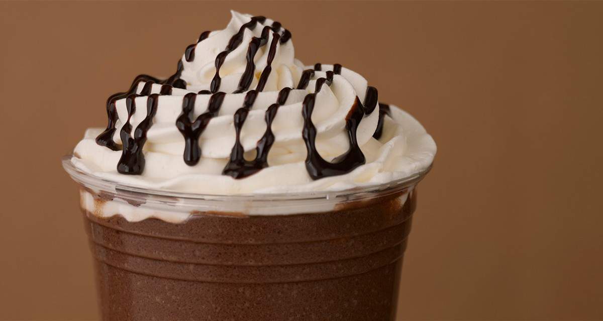 Savor a chocolate milkshake straight out of a Disney park experience, crowned with fluffy whipped cream and drizzled with signature chocolate syrup, all served in a clear plastic cup. The milkshake’s presentation against a rich brown backdrop brings to mind the iconic indulgent desserts found throughout Walt Disney World, capturing the magic of your favorite sweet treat in every detail.