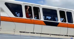A guest waves from the open window of a classic white Disney Monorail accented with an orange stripe, set against a bright blue sky, while fellow riders look on with concern after the monorail unexpectedly loses power during their trip at Walt Disney World.