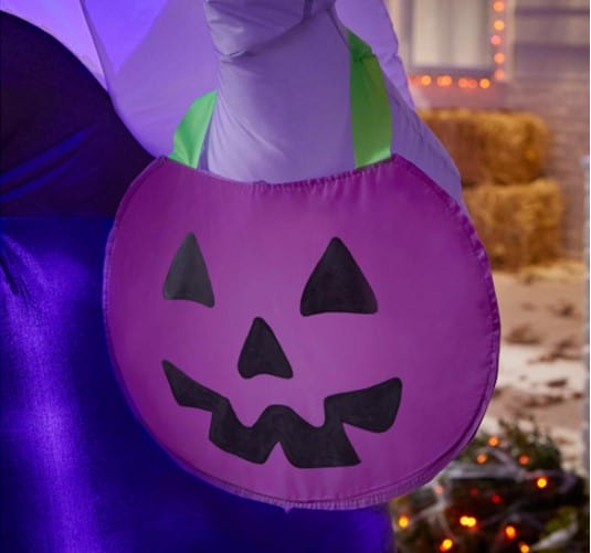 A guest at the theme park holds a vibrant purple pumpkin-shaped bag with a black jack-o'-lantern face, perfect for collecting treats during the park’s Halloween festivities and seasonal décor displays.
