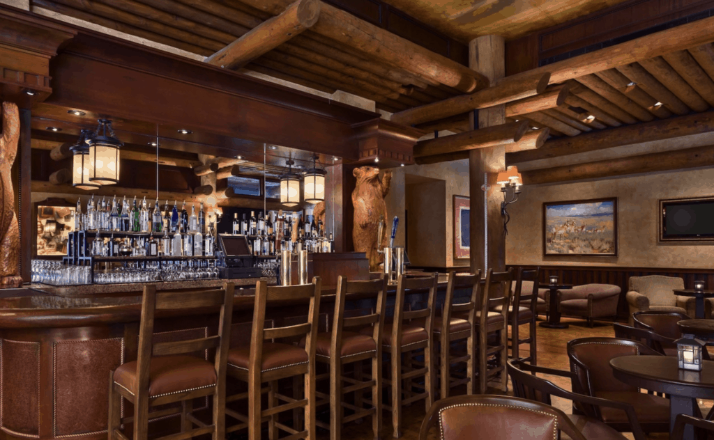 Inviting Wilderness Lodge bar with exposed wooden beams, tall chairs, shelves lined with colorful liquor bottles, and glowing lanterns creating a warm atmosphere—an immersive retreat for Disney guests seeking the ultimate day of relaxation and magic.