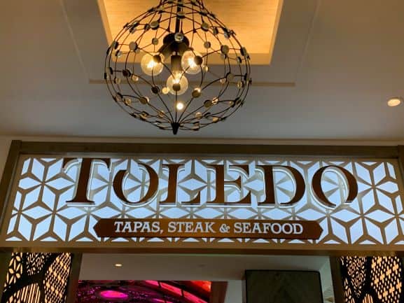 The illuminated "TOLEDO Tapas, Steak & Seafood" sign glows beneath a sleek geometric chandelier, signaling the grand reopening of this beloved Disney dining spot and inviting guests to experience Cast Members' signature hospitality once again.