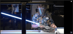 An inventor, shielded for safety, wields a glowing blue real lightsaber at a cluttered workshop bench—an eye-catching tribute to the imaginative inventions and behind-the-scenes magic crafted by Disney Imagineering.