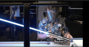 An inventor, shielded for safety, wields a glowing blue real lightsaber at a cluttered workshop bench—an eye-catching tribute to the imaginative inventions and behind-the-scenes magic crafted by Disney Imagineering.