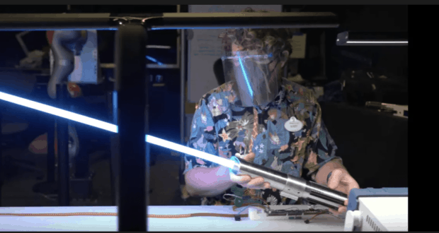 An inventor, shielded for safety, wields a glowing blue real lightsaber at a cluttered workshop bench—an eye-catching tribute to the imaginative inventions and behind-the-scenes magic crafted by Disney Imagineering.