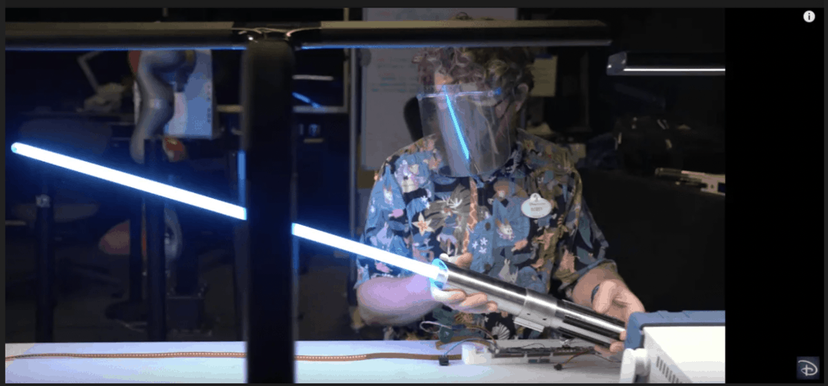 An inventor, shielded for safety, wields a glowing blue real lightsaber at a cluttered workshop bench—an eye-catching tribute to the imaginative inventions and behind-the-scenes magic crafted by Disney Imagineering.