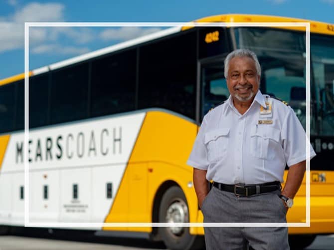A friendly Mears team member welcomes guests in front of a yellow and white Mears Connect bus beneath a clear blue sky, ready to help you book transportation to your Orlando theme park adventure at the latest Magical Express Replacement rates.