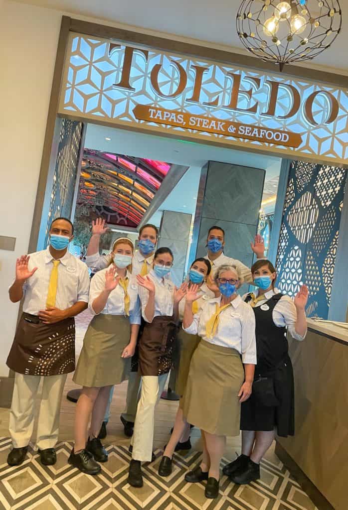 Eight Cast Members from Toledo – Tapas, Steak & Seafood at Disney’s Coronado Springs Resort, dressed in restaurant uniforms and masks, wave beneath the restaurant’s sign as they celebrate the eagerly awaited reopening of this beloved Disney dining spot.