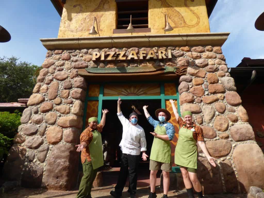 Four Pizzafari Cast Members in their themed uniforms smile together outside the vibrant Pizzafari entrance at Disney’s Animal Kingdom, commemorating the highly anticipated restaurant reopening.