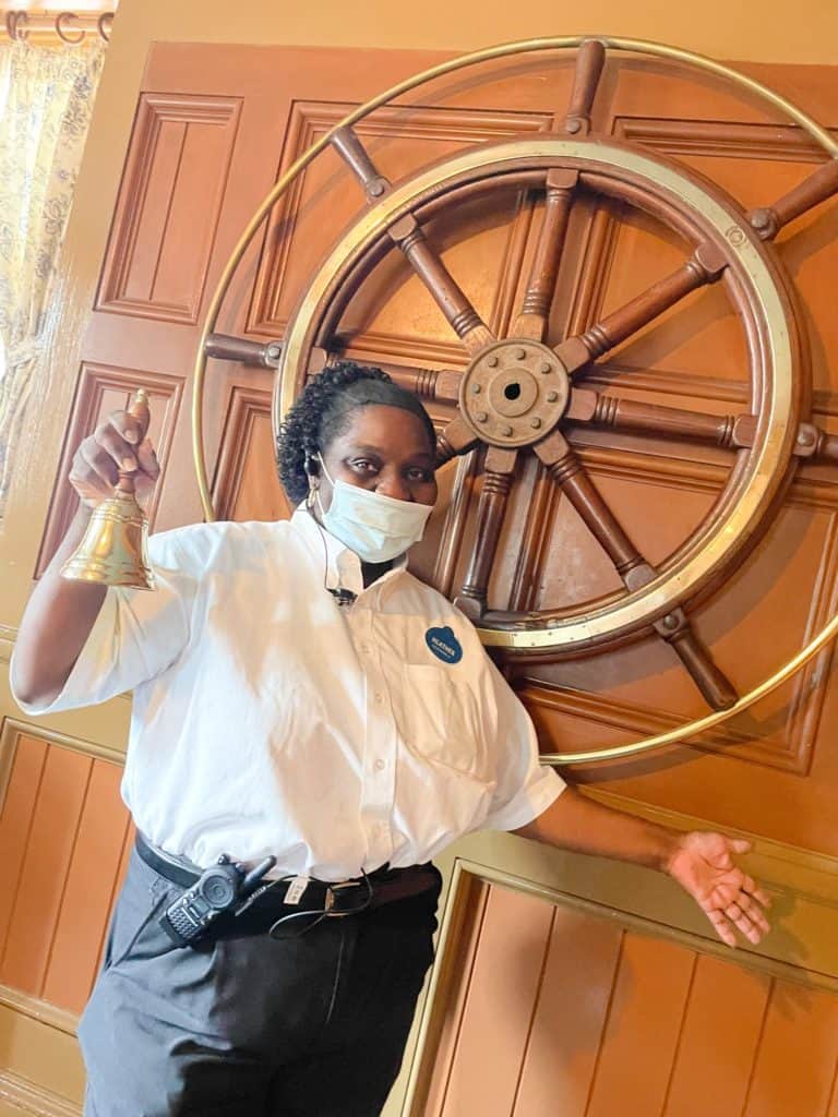 A Disney cast member in themed uniform and mask rings a bell with enthusiasm, standing by a grand ship wheel on the wall, signaling the exciting preparations for beloved Disney restaurants to reopen.