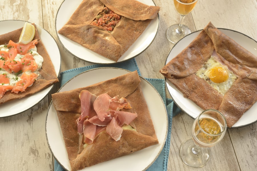 Four savory buckwheat crepes filled with tasty ingredients, artfully arranged on plates alongside glasses of cider atop a rustic wooden table—an authentic treat to enjoy at La Crêperie de Paris in EPCOT’s France Pavilion.