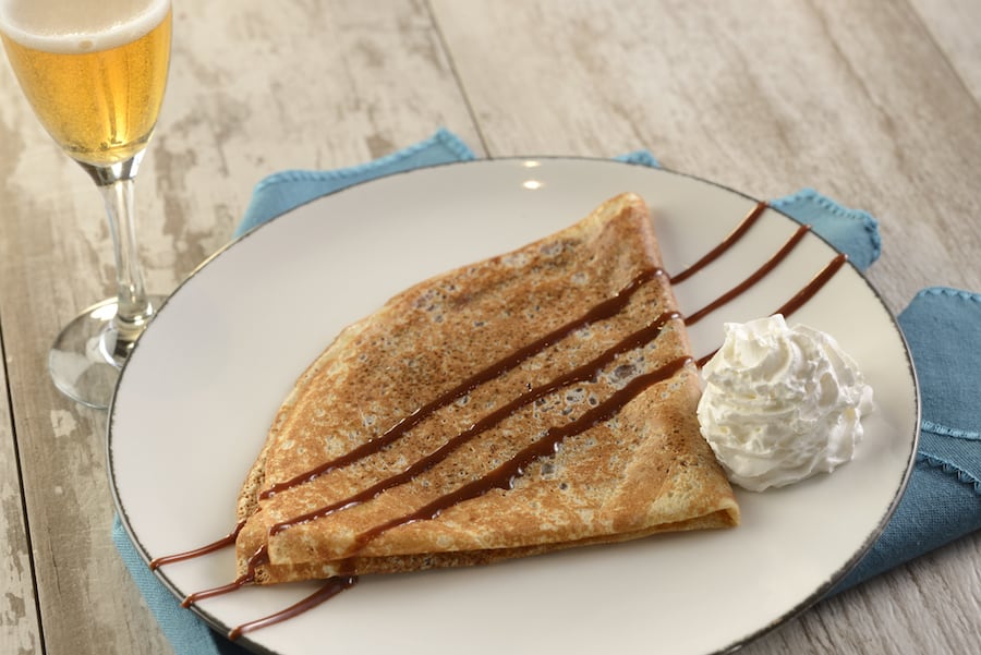A mouthwatering crepe folded and drizzled with chocolate, topped with a swirl of whipped cream on a plate beside a sparkling drink—just like the decadent treats served at La Crêperie de Paris in EPCOT.