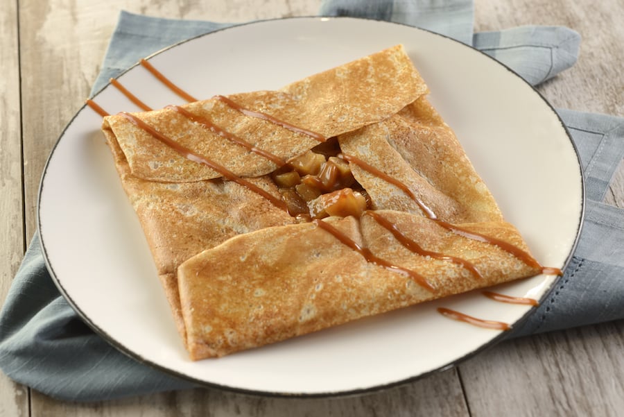A warm, golden crepe folded over sweet diced apples and drizzled with caramel sauce, served on a white plate atop a blue napkin—just like the irresistible desserts guests love at EPCOT’s La Crêperie de Paris.