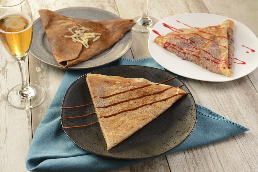 Three folded crepes with assorted toppings are served on plates beside a glass of beer atop a rustic table, creating the mouthwatering look of classic French cuisine found at La Crêperie de Paris in EPCOT—just the kind of delicious treat that makes a day at Walt Disney World Resort even more magical.