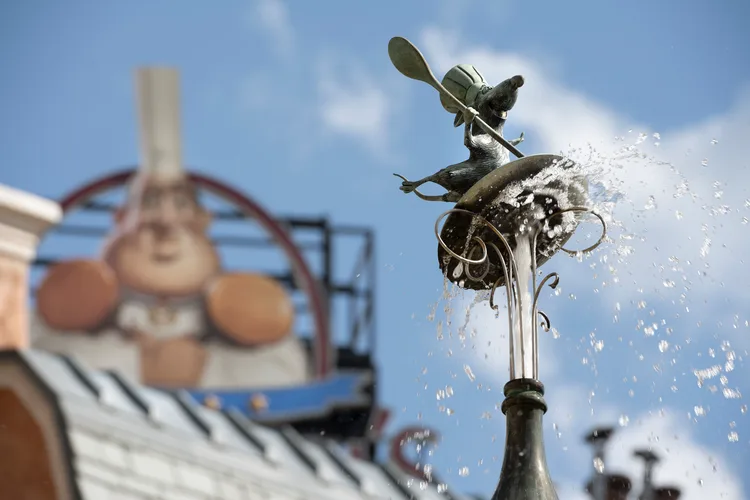 Remy the rat chef statue sprays water from a whimsical fountain, capturing the magic of Remy's Ratatouille Adventure at EPCOT’s France Pavilion, while a playful cartoon chef appears in the softly blurred background.