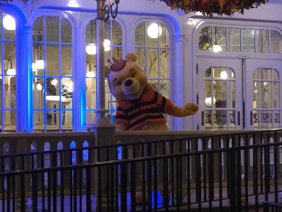 Winnie the Pooh dressed as a bee waves cheerfully in front of a glowing building at Magic Kingdom during Disney After Hours Boo Bash, adding to the Halloween excitement.