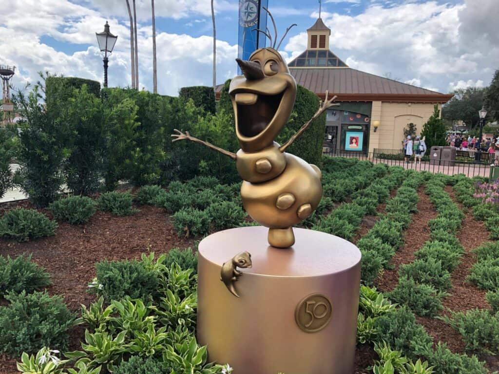 Olaf from Frozen, gleaming in gold with his signature joyful grin and open arms, welcomes guests among lush greenery as part of the magical Fab 50 Character Collection at EPCOT.