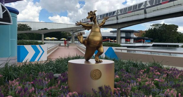 The gleaming gold dragon statue from the Fab 50 Character Collection dazzles in a lively flower garden at EPCOT, set against iconic monorail tracks and dramatic clouds—making it a must-see photo spot for Disney fans.