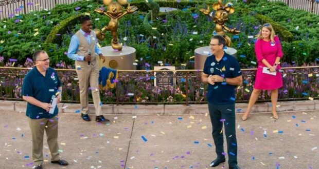 Guests gather beside shimmering gold Mickey and Minnie statues from the Fab 50 Character Collection in a festive park scene at Walt Disney World, with confetti and vibrant flowers adding to the celebration as Cast Members join in the fun nearby.