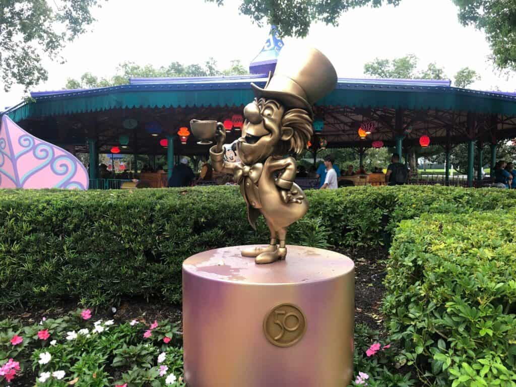 The Mad Hatter’s golden statue raises a teacup in salute, set against the whimsical backdrop of the teacups ride and lush gardens at Magic Kingdom, part of Walt Disney World’s dazzling 50th Anniversary celebration.
