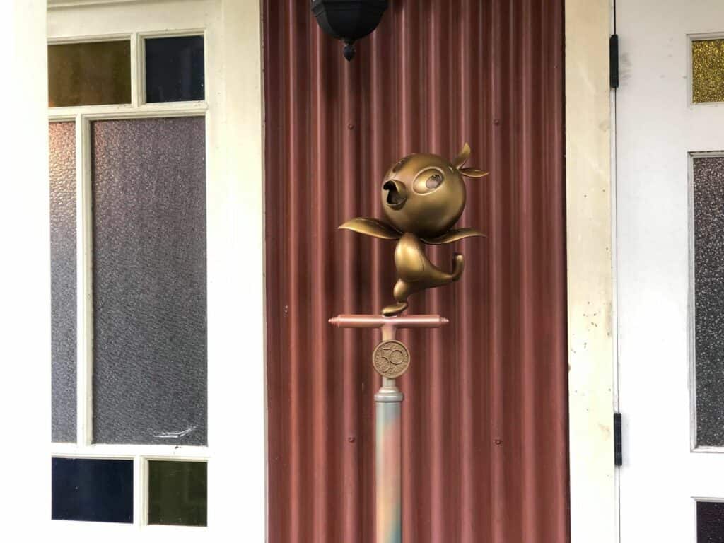 A whimsical bronze statue of a singing cartoon bird perched on a pedestal pops against the bold red corrugated wall and doors, capturing the playful spirit of Magic Kingdom during Walt Disney World’s 50th Anniversary celebration.