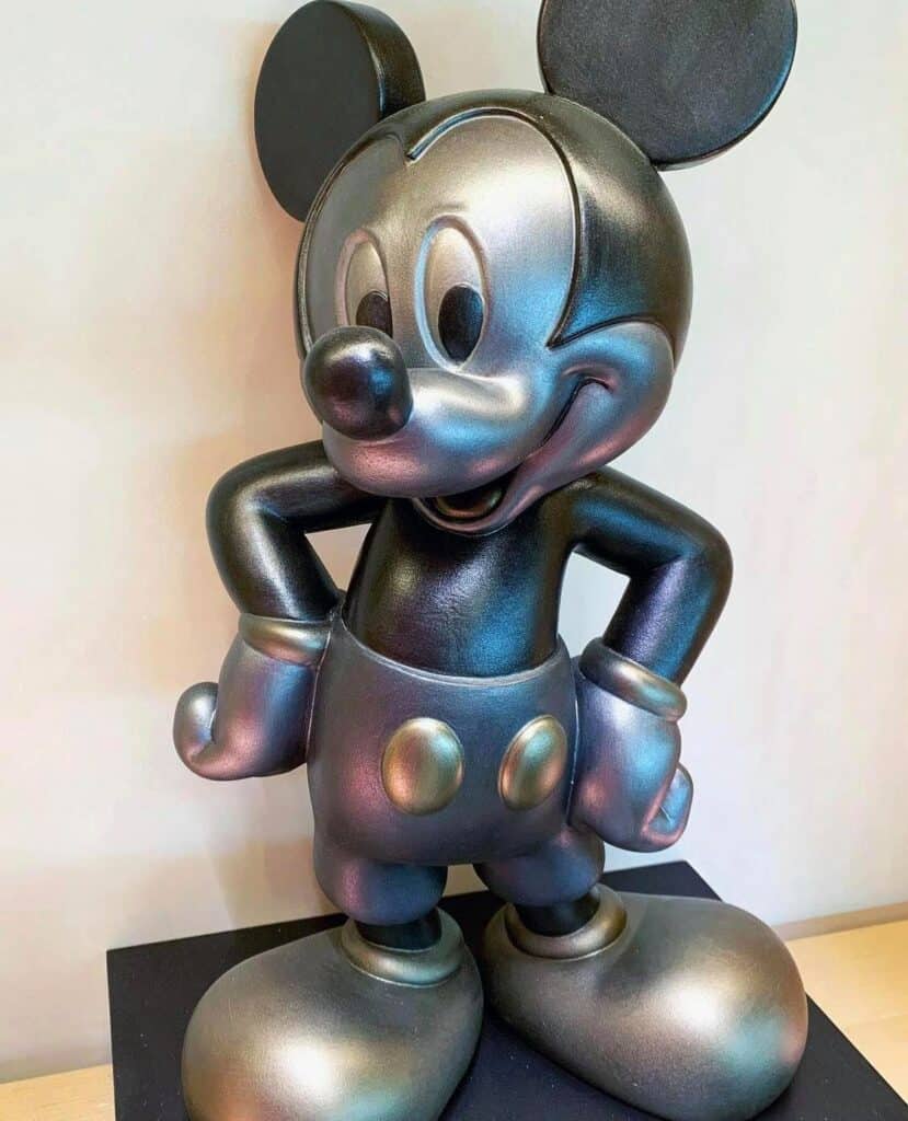 A metallic Mickey Mouse statue, inspired by the beloved Disney icon, stands confidently with hands on hips and faces forward against a bright backdrop—an artistic tribute to Disney creativity on display at Creations Shop in EPCOT.