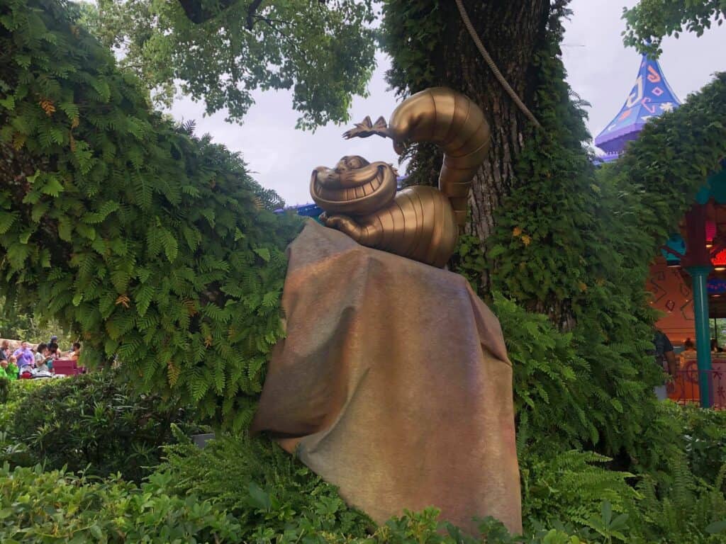 The gold Cheshire Cat statue gleams atop a rock surrounded by lush greenery and trees at Magic Kingdom, part of Walt Disney World's dazzling 50th Anniversary celebration.