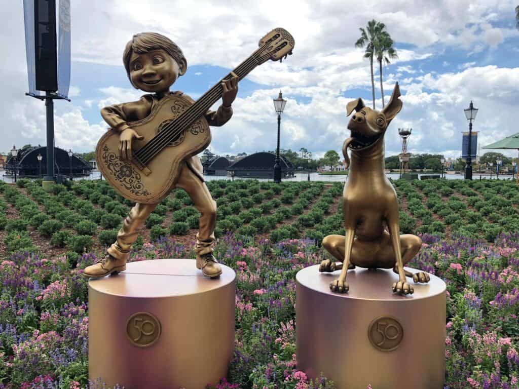 Bronze statues of Miguel strumming his guitar alongside Dante from Coco, displayed on pedestals amid vibrant flowers, celebrate the Fab 50 Character Collection at EPCOT.