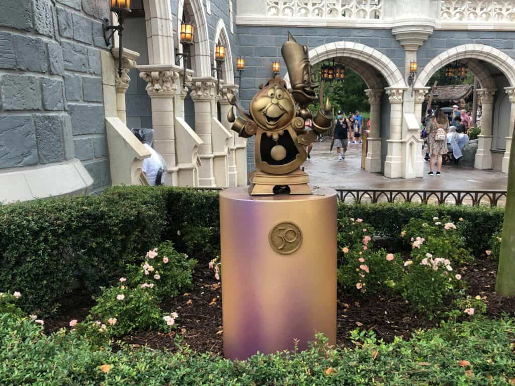 Cogsworth shines in gold on a pedestal surrounded by vibrant flowers and greenery near the castle entrance, commemorating Magic Kingdom’s 50th Anniversary at Walt Disney World.
