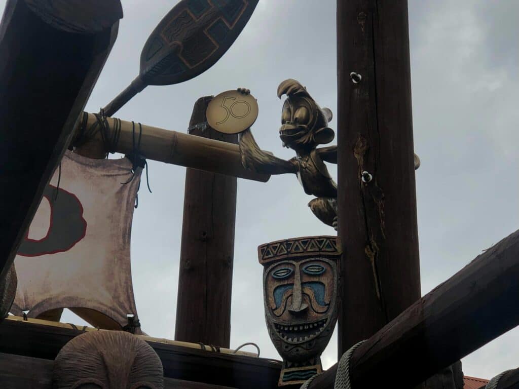 Jose Carioca statue proudly displays a golden "50" medallion atop Adventureland’s wooden beams, with iconic Enchanted Tiki Room tribal masks below, all set against a cloudy sky to mark Magic Kingdom’s 50th Anniversary celebration.