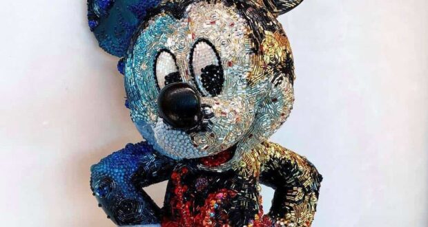 A whimsical beaded Mickey Mouse sculpture featuring his signature blue ear, black nose, and bright red outfit stands out against a plain wall—an eye-catching tribute to Disney magic from Creations Shop.